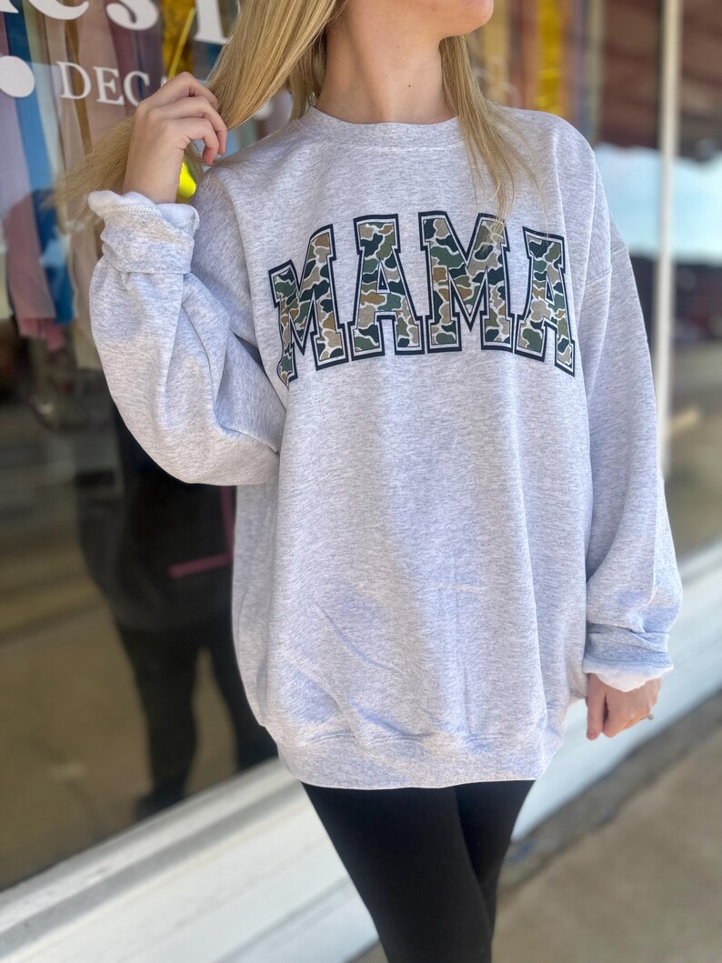 Puede incluir: Una sudadera gris con un gran gr&aacute;fico de "MAMA" en un patr&oacute;n de camuflaje. Las letras est&aacute;n delineadas en negro.