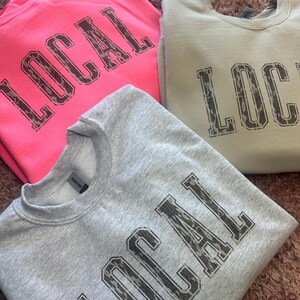 May include: Three sweatshirts with the word "LOCAL" printed in a camouflage pattern. The sweatshirts are in pink, light gray, and light green.