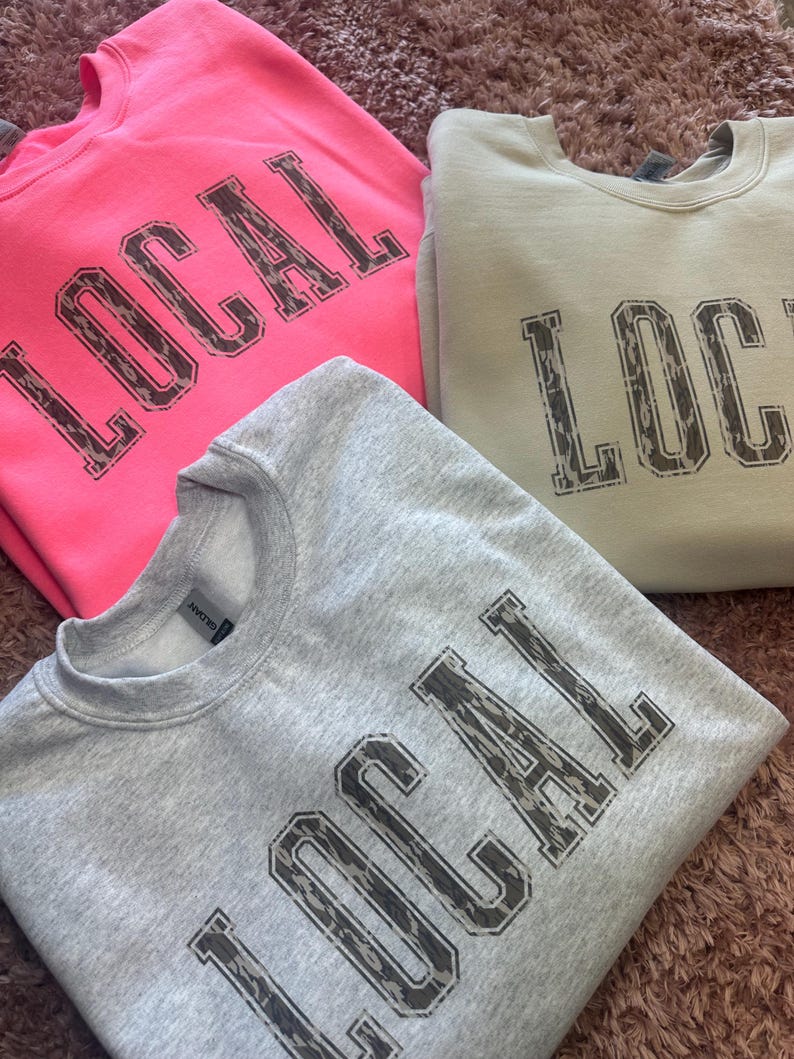 May include: Three sweatshirts with the word "LOCAL" printed in a camouflage pattern. The sweatshirts are in pink, light gray, and light green.