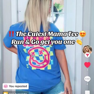 May include: A woman wearing a blue t-shirt with a pink and green smiley face graphic and the text "The Cutest Mama Tee" on it. She is also wearing denim shorts.