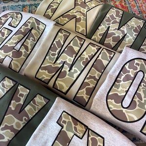 May include: Five different shirts with camouflage print letters. The letters spell out "TEXAS" in a large font. The shirts are in various shades of gray and green.