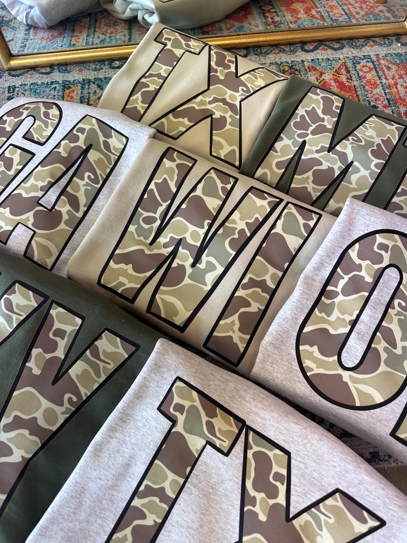 May include: Five different shirts with camouflage print letters. The letters spell out "TEXAS" in a large font. The shirts are in various shades of gray and green.