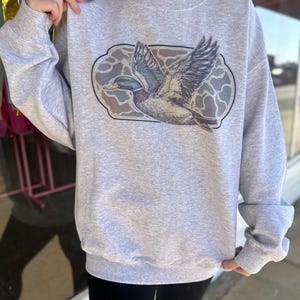 May include: A gray sweatshirt with a graphic of a flying duck in a camouflage pattern.