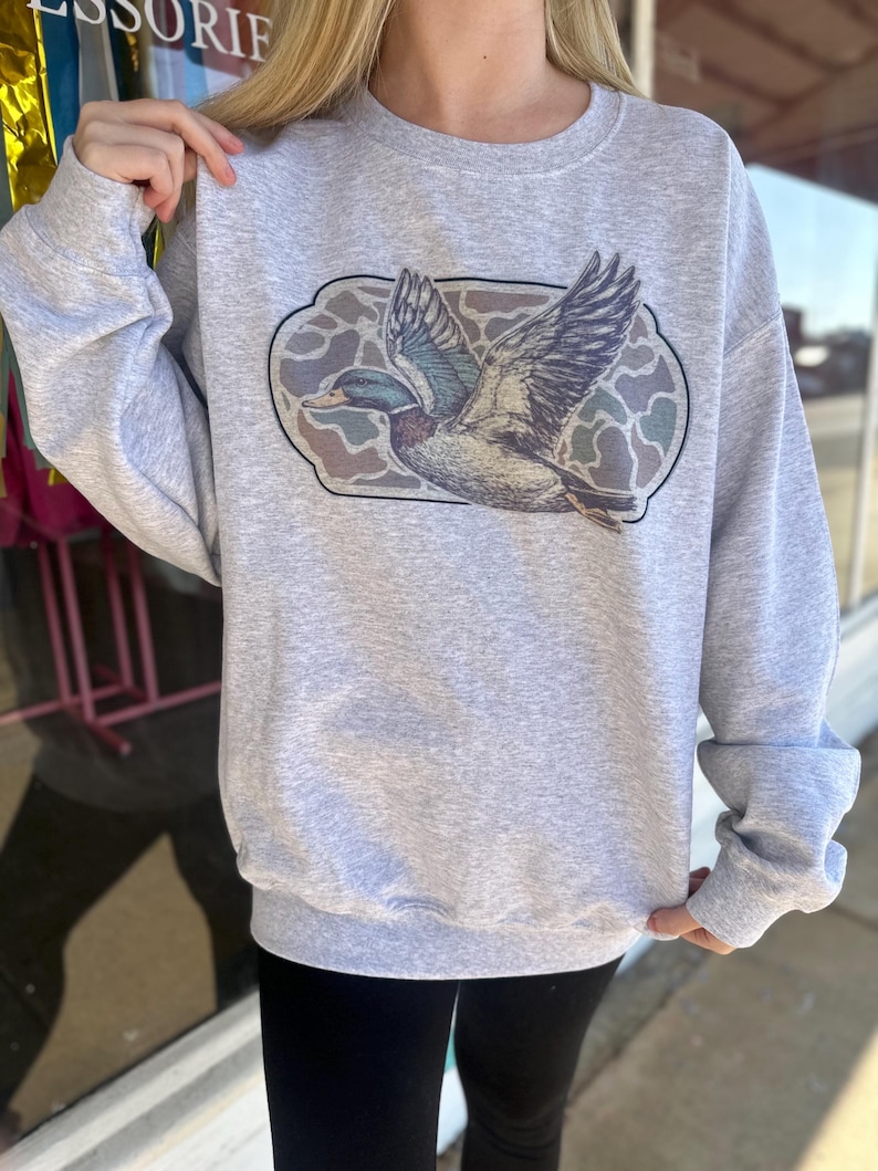 May include: A gray sweatshirt with a graphic of a flying duck in a camouflage pattern.