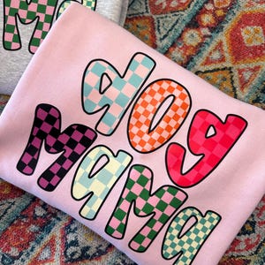 May include: Pink sweatshirt with the words "dog mama" in colorful checkered letters.