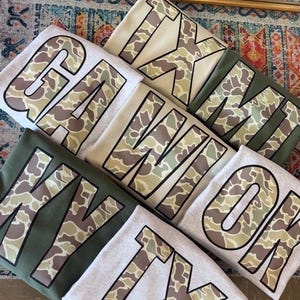 May include: A stack of five gray, beige, and green t-shirts with camouflage print letters spelling out "GA, WI, OK, TX, TX".