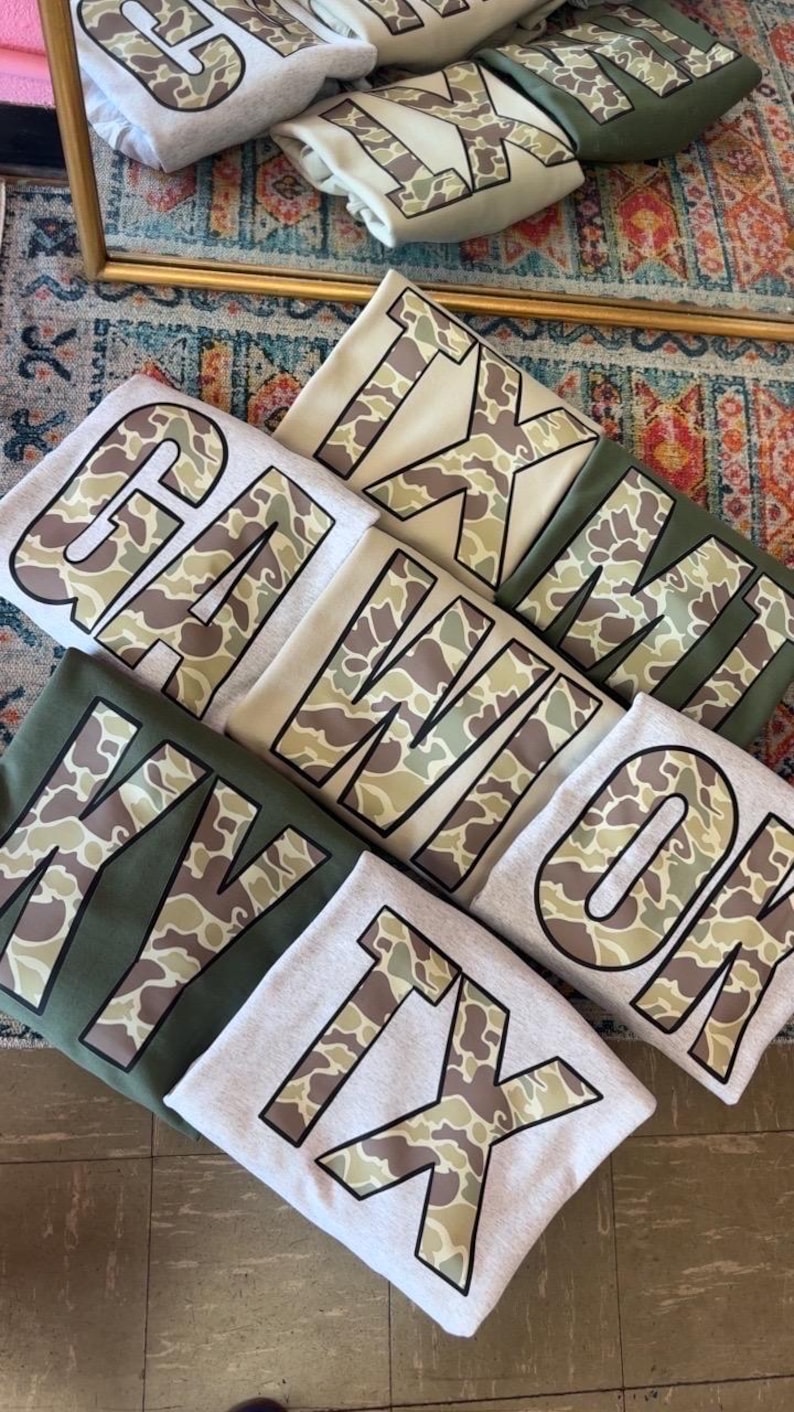 May include: A stack of five gray, beige, and green t-shirts with camouflage print letters spelling out "GA, WI, OK, TX, TX".