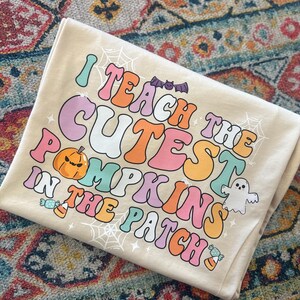 May include: A beige t-shirt with the text "I teach the cutest pumpkins in the patch" in rainbow colors. The text is in a retro font style. There are also images of a pumpkin, a ghost, and candy corn.