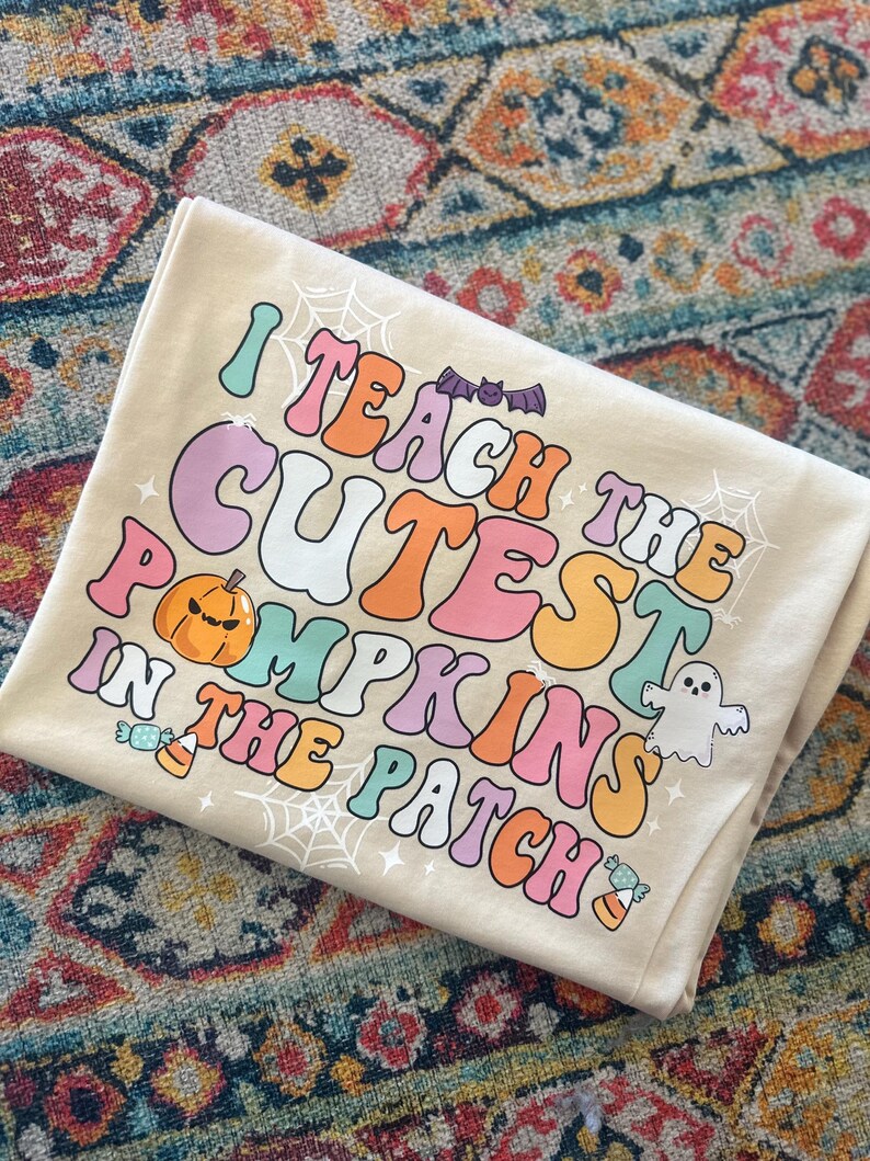 May include: A beige t-shirt with the text "I teach the cutest pumpkins in the patch" in rainbow colors. The text is in a retro font style. There are also images of a pumpkin, a ghost, and candy corn.