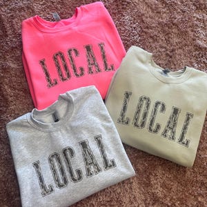 May include: Three sweatshirts with the word "LOCAL" printed on them in a leopard print font. The sweatshirts are in pink, gray, and beige.