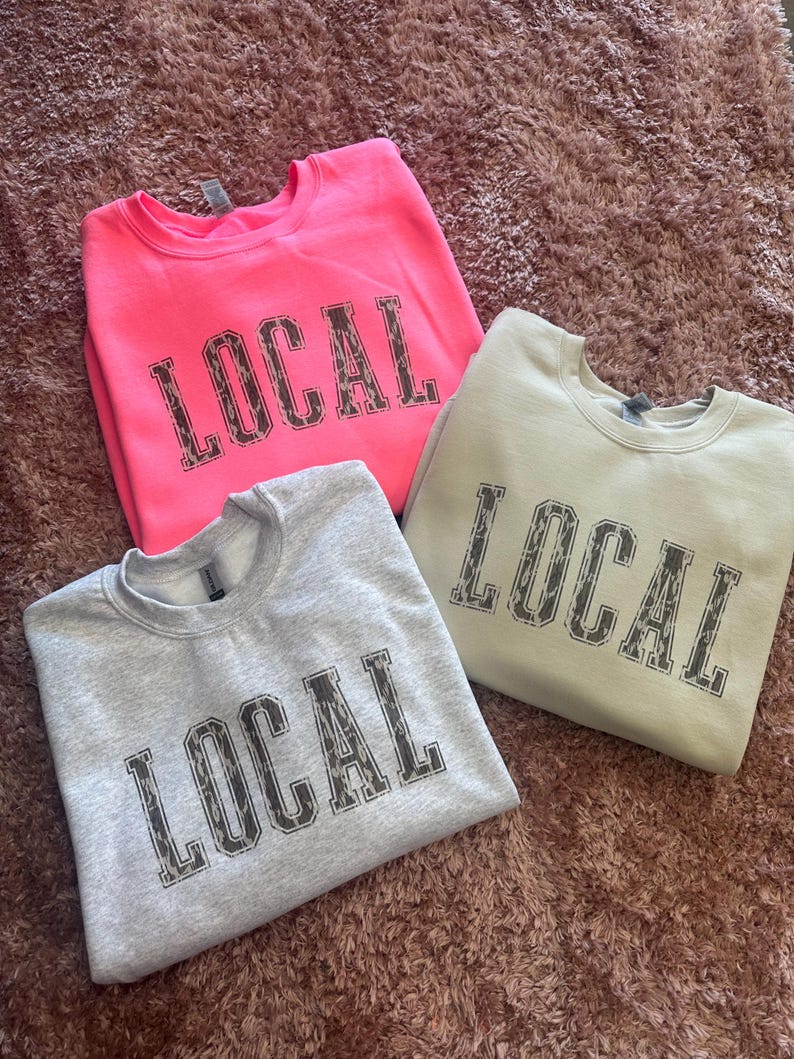 May include: Three sweatshirts with the word "LOCAL" printed on them in a leopard print font. The sweatshirts are in pink, gray, and beige.