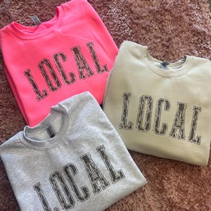May include: Three sweatshirts with the word "LOCAL" printed in a leopard print design. One sweatshirt is pink, one is light gray, and one is beige.