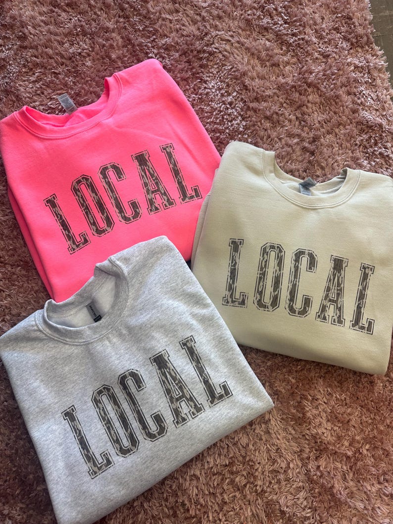 May include: Three sweatshirts with the word "LOCAL" printed in a leopard print design. One sweatshirt is pink, one is light gray, and one is beige.