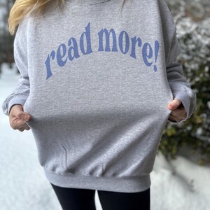 Puede incluir: Una sudadera gris con el texto "read more!" en letras azules.
