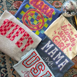 May include: Five different t-shirts with various designs and text. The shirts are all folded and stacked on top of each other. The top shirt is blue with a yellow and green checkered pattern and a pink smiley face with the text "MAMA" in pink. The second shirt is gray with the text "WIFEY" in pink and white. The third shirt is cream with the text "I'm literally just a girl" in pink. The fourth shirt is blue with the text "MAMA" in purple. The bottom shirt is light blue with the text "USA" in red and white stars.