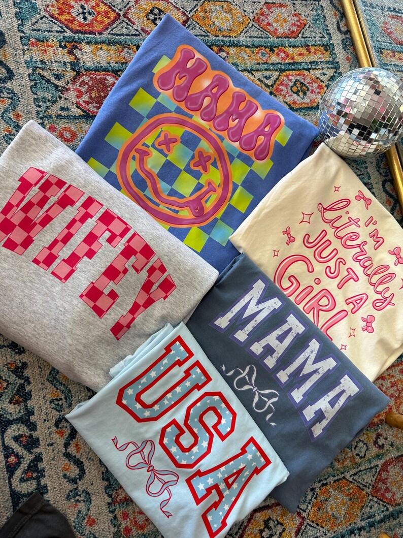 May include: Five different t-shirts with various designs and text. The shirts are all folded and stacked on top of each other. The top shirt is blue with a yellow and green checkered pattern and a pink smiley face with the text "MAMA" in pink. The second shirt is gray with the text "WIFEY" in pink and white. The third shirt is cream with the text "I'm literally just a girl" in pink. The fourth shirt is blue with the text "MAMA" in purple. The bottom shirt is light blue with the text "USA" in red and white stars.