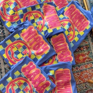 May include: A stack of blue t-shirts with a checkered pattern and a smiley face graphic. The shirts have different text on them, including "Happy Hour", "Witty", "SAMA", and "Dog Mom".