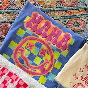 May include: A blue t-shirt with a checkered pattern and a pink smiley face with X's for eyes. The word "MAMA" is written in pink bubble letters above the smiley face.
