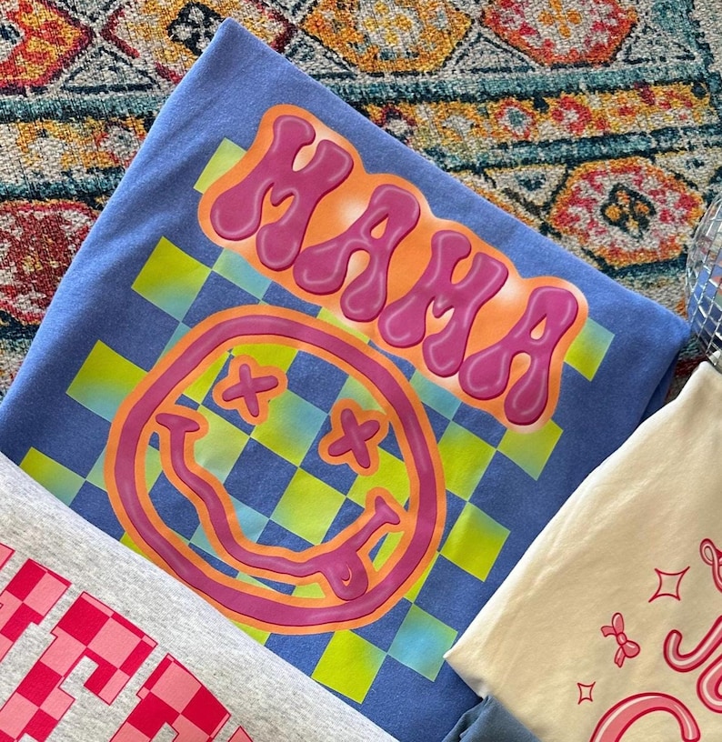 May include: A blue t-shirt with a checkered pattern and a pink smiley face with X's for eyes. The word "MAMA" is written in pink bubble letters above the smiley face.