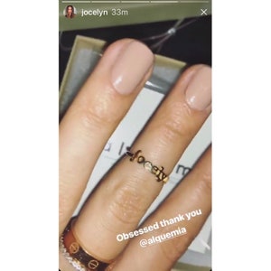 May include: A close-up of a hand wearing a gold ring with the word "Hope" spelled out in small letters. The ring is on the ring finger of the left hand. The hand is also wearing a silver band ring on the little finger. The caption reads "Obsessed thank you @alquemia".