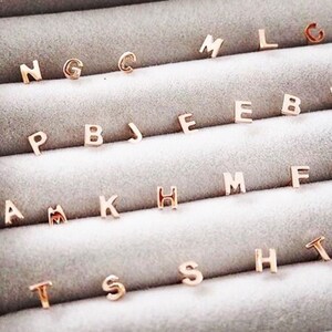 May include: A close-up of a display of gold-toned initial letter charms. The charms are arranged in rows, with each row containing a different letter of the alphabet. The charms are small and delicate, and they would make a great addition to any jewellery collection.