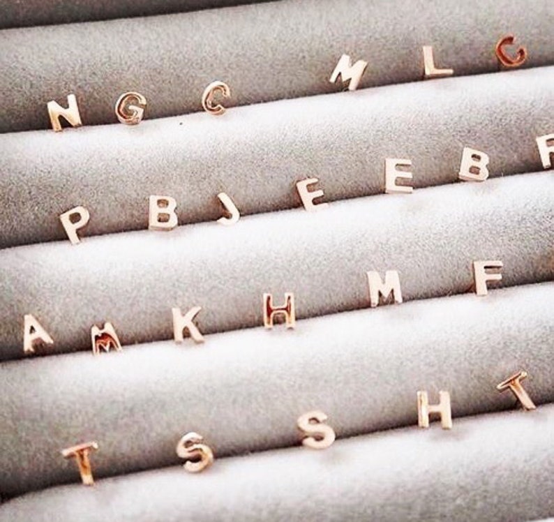 May include: A close-up of a display of gold-toned initial letter charms. The charms are arranged in rows, with each row containing a different letter of the alphabet. The charms are small and delicate, and they would make a great addition to any jewellery collection.