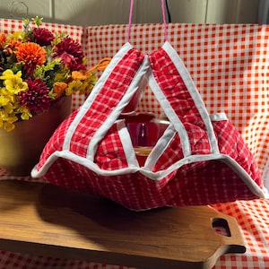May include: A quilted fabric bowl cozy featuring a red and white checkered pattern with white fabric trim along the edges. The cozy has four triangular flaps that meet at the top, held together by a thin pink string for hanging display.