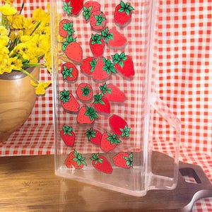 May include: A clear acrylic pitcher featuring a repeating pattern of red strawberries with green leaves. The rectangular container has a sturdy handle on the right side and a flat base, designed for serving beverages in a kitchen or dining setting.