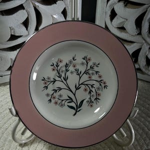 Peut inclure: Une assiette vintage rose et blanche avec un motif floral. L'assiette a un bord rose avec un bord noir et un centre blanc. Le centre de l'assiette présente un motif d'arbre noir avec des fleurs roses et grises. L'assiette est présentée sur un support en métal blanc.