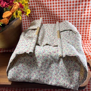 May include: A fabric basket with a cream background featuring a small floral pattern in pink, blue, and green. The basket has two light-colored wooden dowel handles attached to the top edges, allowing the fabric to drape between them.