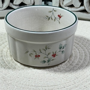 May include: A white ceramic bowl with a green rim and a floral design featuring green leaves and red berries. The bowl has a round shape and is sitting on a white, textured surface.
