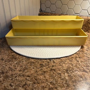 May include: Two yellow plastic stackable storage containers with scalloped edges. The containers are empty and sitting on a brown countertop.