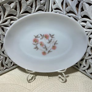 May include: Oval, white serving dish with a floral design in the center. The flowers are pink with brown stems and leaves. The dish is displayed on a white metal stand. A decorative piece for serving food.