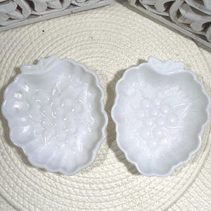 Può includere: Due piatti bianchi a forma di foglia con un motivo a uva. I piatti sono realizzati in materiale lucido e poggiano su una superficie intrecciata color crema. Intagli in legno decorativi sullo sfondo.