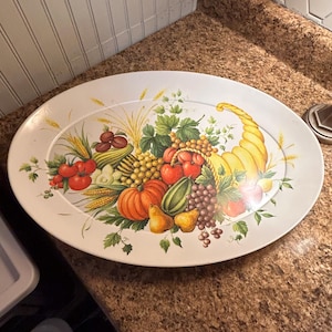 Puede incluir: Un plato de servir ovalado blanco con un diseño de cornucopia que presenta una variedad de frutas y verduras, incluyendo uvas, peras, calabazas y tomates.