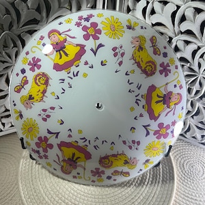 May include: A white, round ceiling light fixture with a whimsical design. The fixture features illustrations of a girl in a yellow dress, yellow sheep, flowers, and butterflies in shades of pink, yellow, and purple. The fixture is supported by a black metal frame.