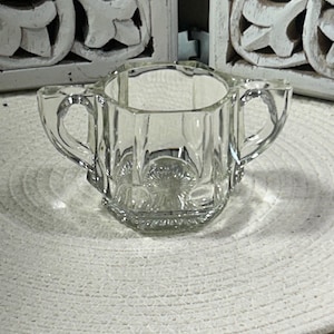 以下が含まれることがあります： 2つのハンドルが付いた、透明なヴィンテージガラスの砂糖入れ。ボウルはファセットデザインで、丸いテクスチャのある表面に置かれています。砂糖入れは空で、状態は良好に見えます。背景には、白い彫刻が施された木製パネルが含まれています。