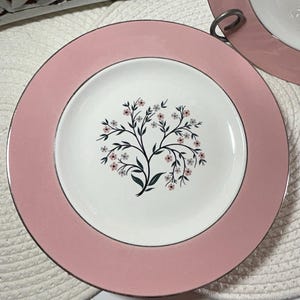 Puede incluir: Un juego de platos de cerámica rosa y blanco. Los platos tienen un borde rosa y un centro blanco con un diseño de árbol negro y pequeñas flores rosas. Los platos tienen un ribete plateado.