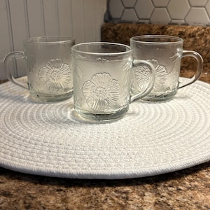 May include: Three clear glass mugs with a floral design. The mugs have handles and are sitting on a white round placemat.
