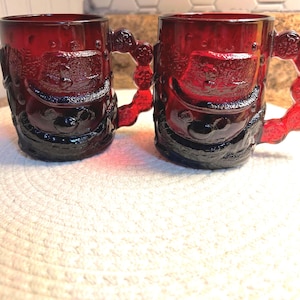 Op de afbeelding: Twee robijnrode glazen mokken met reliëfontwerpen van de kerstman. De mokken hebben gedetailleerde handvatten en bodems, en het kerstmanontwerp is zichtbaar op de voorkant van elke mok. De mokken staan op een wit oppervlak.