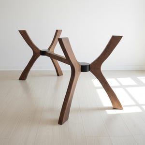 Puede incluir: Dos bases de mesa de madera con un dise&ntilde;o &uacute;nico y moderno. Las bases son de madera marr&oacute;n oscuro y presentan un dise&ntilde;o de patas curvadas en forma de X. Cada base tiene un conector circular negro en el centro. Las bases est&aacute;n sobre una pared blanca y un suelo claro.