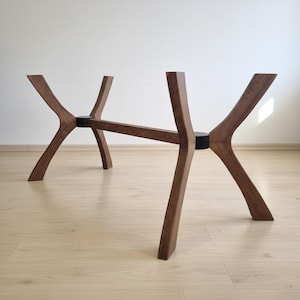 May include: A wooden table base with a unique, modern design. The base is made of dark brown wood and features four angled legs connected by a horizontal beam. The legs are joined with black circular connectors. The table base is designed to support a tabletop.