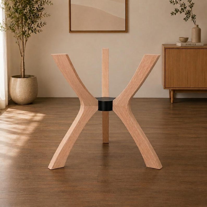 May include: A table base featuring four light-toned wooden legs arranged in an X-shape configuration. The legs are connected at the center by a cylindrical black metal hub, providing a stable foundation for a tabletop that is not currently attached.