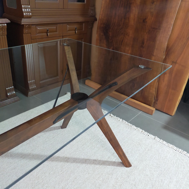 May include: A glass-top table with a wooden base. The base is made of four pieces of wood that are joined together in the center. The table is on a white rug.