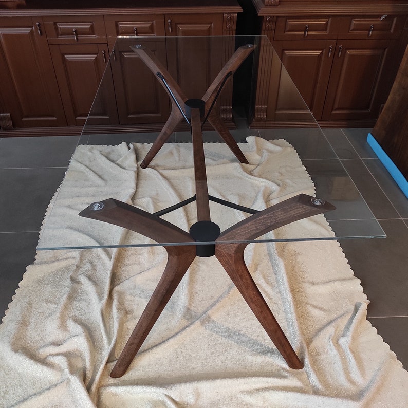 May include: A glass-top dining table with a unique, modern design. The table features a dark wood base with a criss-cross pattern and four legs that extend outward.