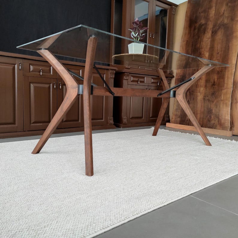 May include: A glass-top dining table with a wooden base. The base is made of dark wood and has a unique X-shaped design. The table is sitting on a white rug.