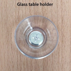 Puede incluir: Un soporte de mesa de vidrio transparente y circular con un tornillo met&aacute;lico en el centro. El texto "Glass table holder" est&aacute; impreso encima del soporte. El soporte est&aacute; sobre una superficie de madera marr&oacute;n claro.
