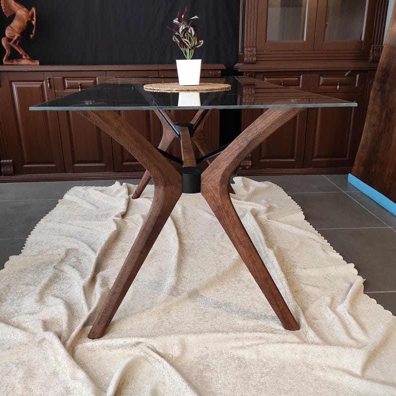 May include: A glass-top dining table with a unique, modern design. The table has a dark wood base with four legs that are shaped like an X. The table is sitting on a white rug.