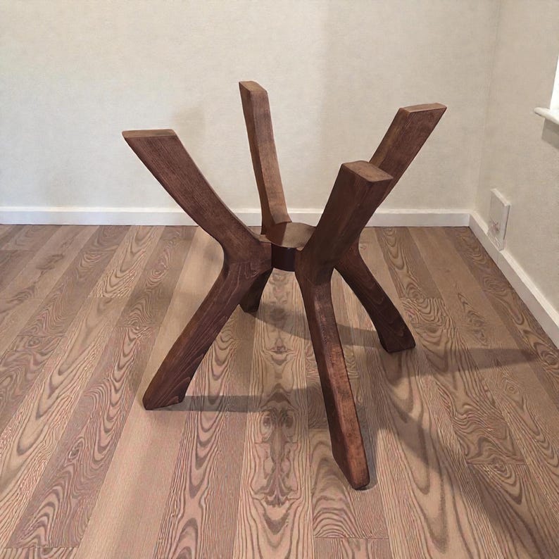 May include: A dark brown wooden table base featuring four angled, splayed legs that converge at a central circular hub. The structure is crafted from solid wood with a visible grain pattern, providing a sturdy foundation for a tabletop surface.