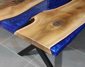 Puede incluir: Una mesa de madera con una incrustación de resina epoxi azul. La mesa tiene un acabado de madera natural y un diseño de borde vivo. La mesa está soportada por una base de metal negro en forma de X.